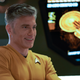 Pike, in his orange captain's uniform, smiles in front of a computer display of the Enterprise.